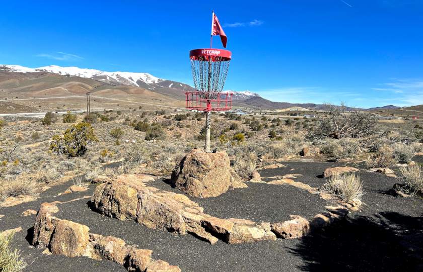 a disc golf basket on top of a rock pyramid surrounded by black cinder-sand with snow-topped mountains in the background.