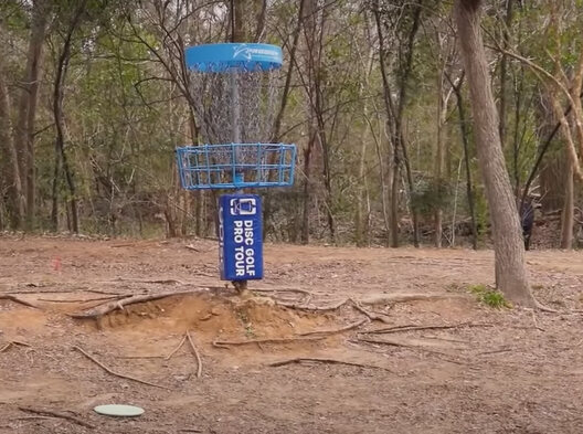 disc golf basket surrounded by bare, hard soil and exposed tree roots